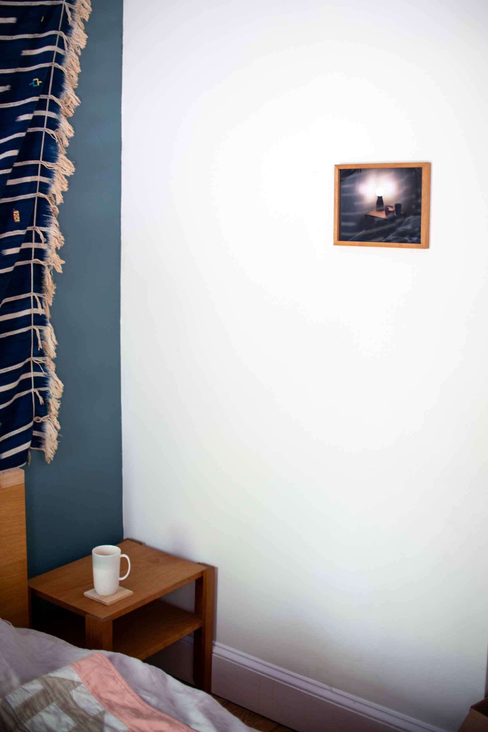 the framed print on the wall with the pictured nightstand in the background along with a bed and a wall hanging textile.